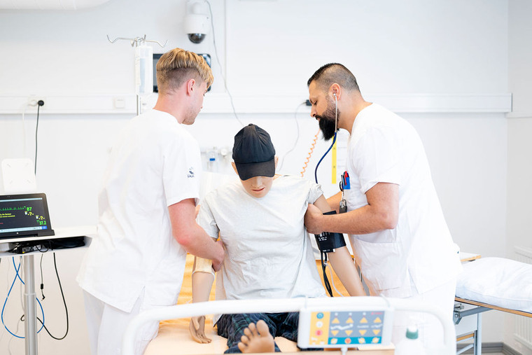 Två sjuksköterskestudenter arbetar med en simuleringsdocka som sitter i en sjukhussäng.