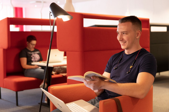 Leende student med lärobok och laptop på bibliotek.
