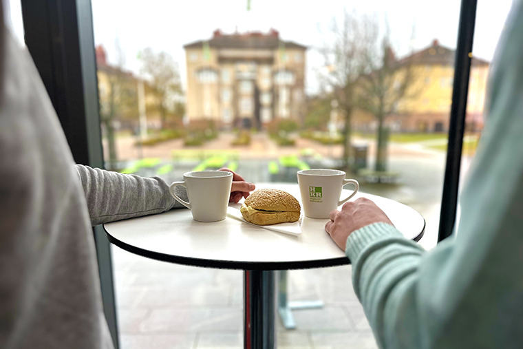 Två personer står vid ett bord med kaffe och macka.