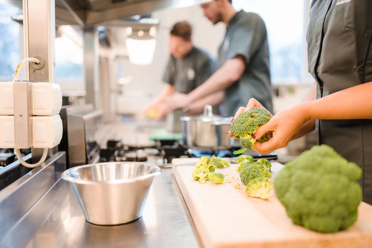 Tre personer i kök, personen i förgrunden delar broccoli.