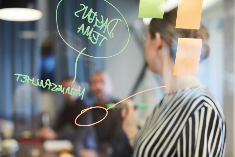 Färgade post-it-lappar med handskriven text fästa på en glasvägg, med affärsklädda personer i bakgrunden.