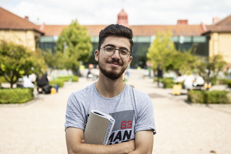 Manlig student står med händerna i kors mitt på campus med glashuset i bakgrunden.