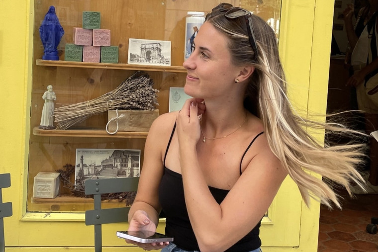 Alicia under en utlandsresa, leende med mobil i handen framför en gul hylla.