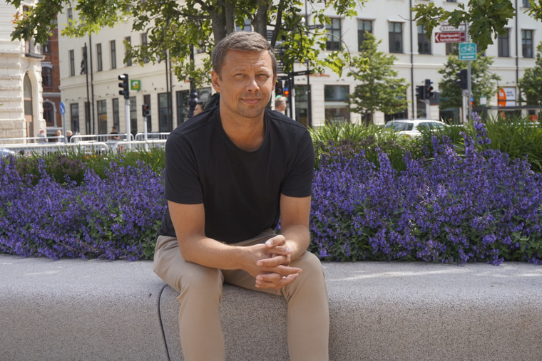 Timmy Sjö sittandes på en stenbänk kantad av lila blommor i stadsmiljö.
