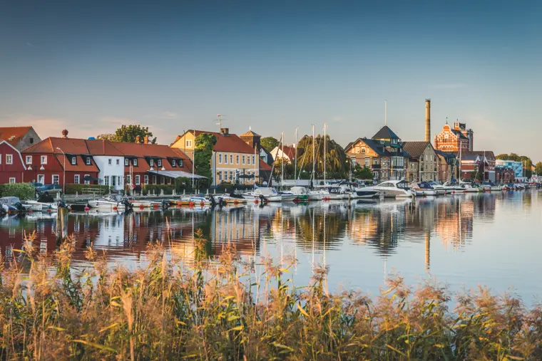 Sm&aring;b&aring;tshamn med stilla vatten och hus i bakgrunden.