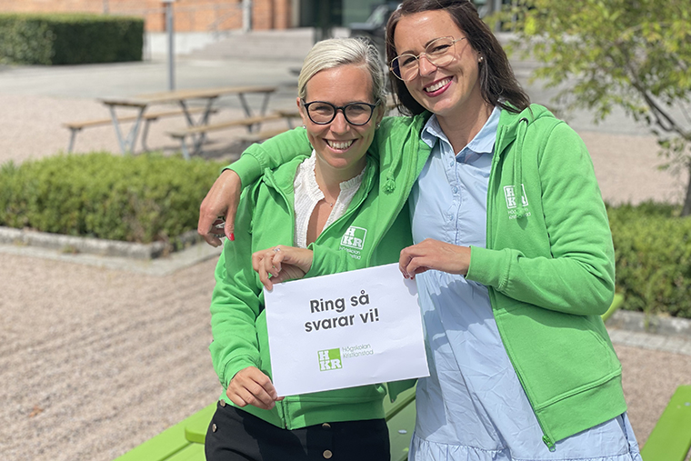 Programledarna för Ring så svarar vi står på campus och håller i en skylt där det står "Ring så svarar vi"