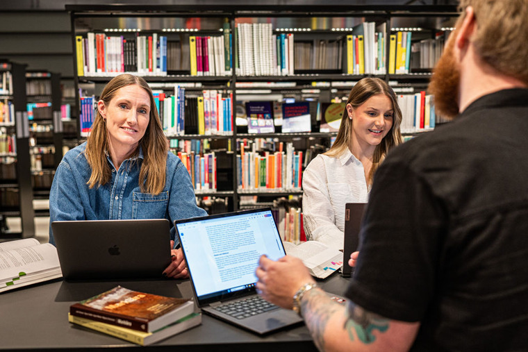 Tre studenter i biblioteket, fokuserade på studier vid ett bord med anteckningar och laptops.