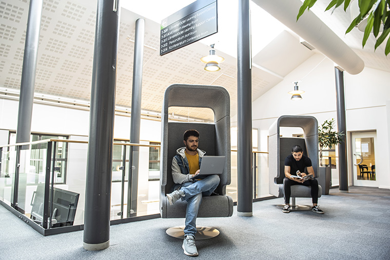 Två studenter sitter i fåtöljer med dator och bok.